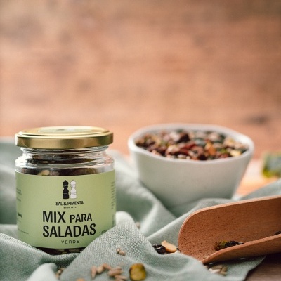 Frasco de mix para saladas verde, colher de madeira e taça branca com mistura, sobre pano verde claro.