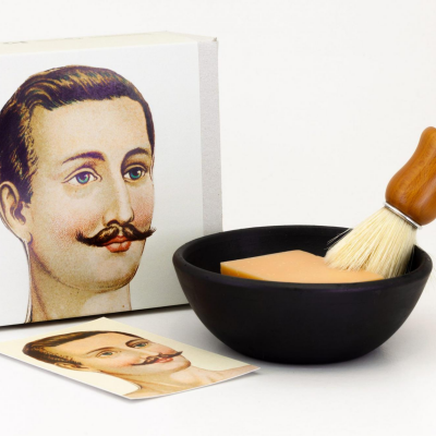 Conjunto de barbear com tigela preta, sabonete salmão, pincel cabo madeira e caixa com imagem de homem com bigode.