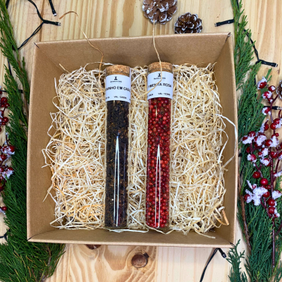 Caixa com tubos de vidro contendo pimenta preta em grão e pimenta rosa, sobre palha decorativa em caixa de cartão com decoração natural