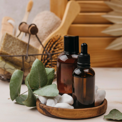 Frascos de vidro castanhos com pedras brancas e folhas verdes em base de madeira, utensílios de banho ao fundo