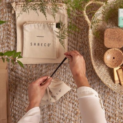 Sacos de tecido cru com texto SHAEO e objetos de cortiça e ráfia numa superfície texturada.