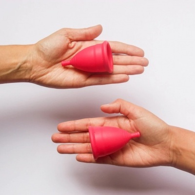 Duas mãos seguram copos menstruais vermelhos de silicone, fundo branco