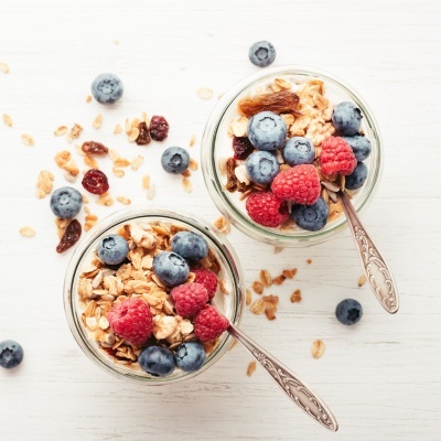 Dois copos de vidro com granola, mirtilos e framboesas sobre mesa branca