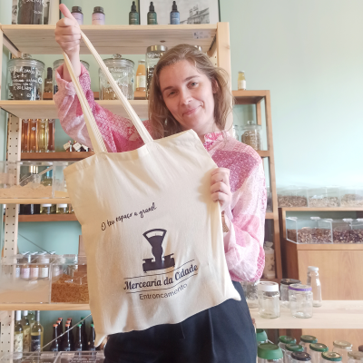 Saco de tecido branco com impressão preta com texto e logótipo, segurado por mulher em loja