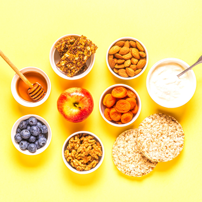 Alimentos diversos saudáveis sobre fundo amarelo com mel, barras de granola, frutos secos, iogurte e maçã