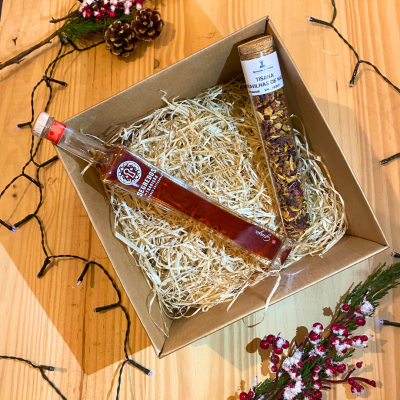 Caixa de presente com garrafa de licor artesanal e tubo de tisana com ervas secas em palha dentro sobre mesa de madeira com decoração natalícia.