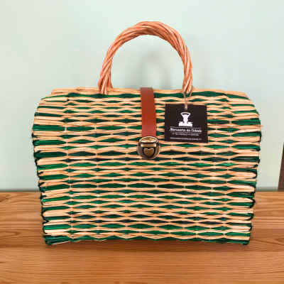 Mala de palha com padrão verde e natural, alça laranja e fecho em coração dourado.