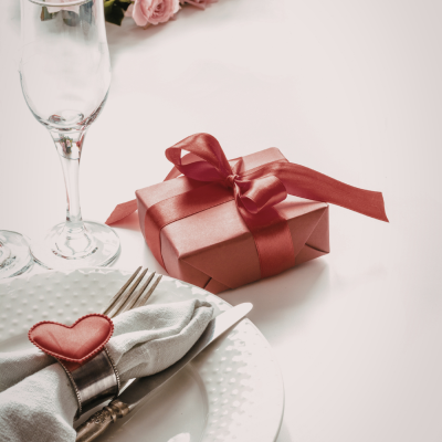 Mesa posta com prato branco, talheres, guardanapo com argola e coração, copo e caixa de presente rosa com fita vermelha.