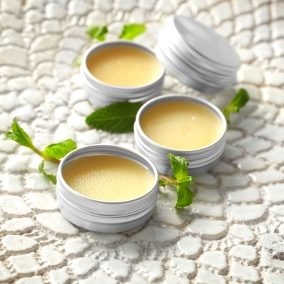 Três frascos pequenos de creme amarelo com tampas prateadas e folhas verdes numa superfície branca texturizada