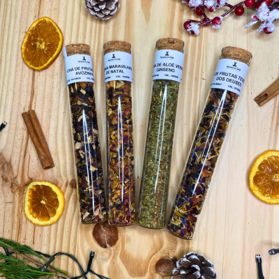 Quatro tubos de vidro transparentes contendo diferentes tipos de chá com etiquetas brancas, sobre madeira com decoração natalícia