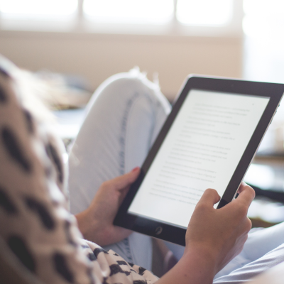 Pessoa segurando tablet preto com ecrã iluminado, vestindo calças de ganga claras e camisa estampada em ambiente interior
