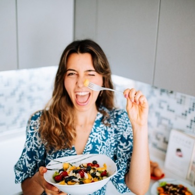 Mulher segura tigela de frutas e iogurte na cozinha, vestindo camisa azul com padrão branco