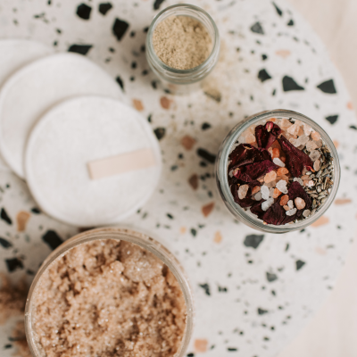 Frascos de vidro com sal grosso e pétalas secas e mistura granulada castanha sobre mesa branca estampada.