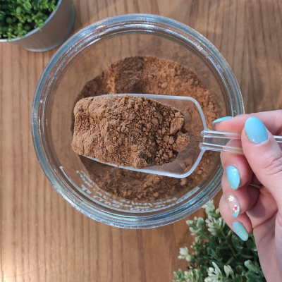 Colher de plástico com pó castanho sobre recipiente de vidro com pó castanho, mãos com unhas azuis decoradas, mesa de madeira e plantas ao fundo