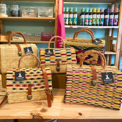 Sacos trançados de palha coloridos com alças de couro numa mesa de madeira em loja.