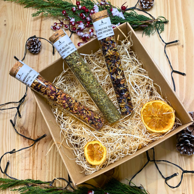 Caixa de presente com três tubos de vidro com ervas e flores secas, decorada com fatias de laranja e ramos verdes com pinhas