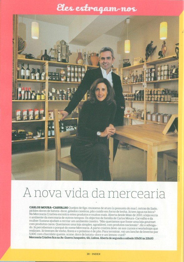 Interior de mercearia com prateleiras cheias e duas pessoas posando perto de uma mesa de madeira.