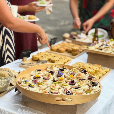 Mesa com mini tostas com figos e flores, quiches pequenas douradas e pessoas a servir-se numa festa exterior.