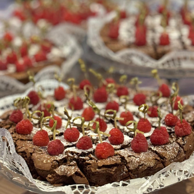 Bolo de chocolate com framboesas e fitas douradas em fatias, sobre papel rendado branco.