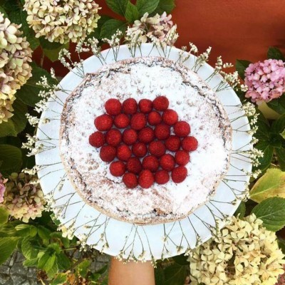 Tarte com açúcar em pó e framboesas sobre um prato branco com borda dourada, rodeada por flores hortênsias.