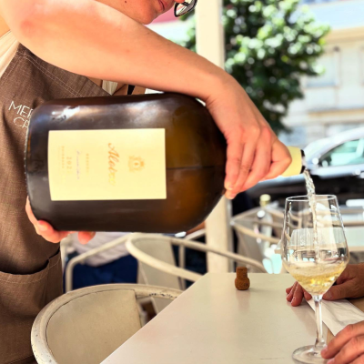 Pessoa a servir vinho branco de garrafa castanha para copo de vidro num café ao ar livre