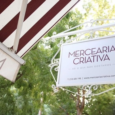 Placa com texto MERCEARIA CRIATIVA num quadro branco decorado, junto a toldo riscado vermelho e branco, com árvores atrás.