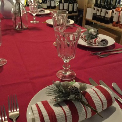 Mesa de jantar posta com decoração natalina, guardanapos listrados e pinhas, garrafas de vinho ao fundo