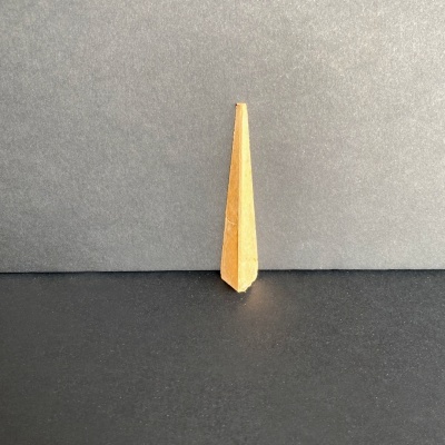 Objeto cónico quadrangular de madeira clara sobre superfície preta e fundo cinzento.