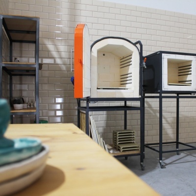 Dois fornos cerâmicos com suportes de metal numa sala com paredes de azulejos brancos e mesa de madeira clara