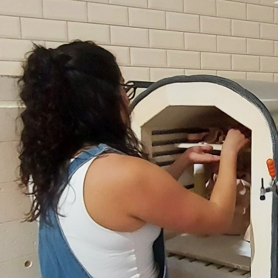 Pessoa usando camisola branca e azul a manusear forno de cerâmica branco junto a parede de azulejos brancos