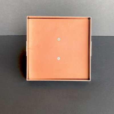 Caixa quadrada castanho alaranjada com dois parafusos prateados em superfície preta