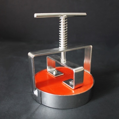 Prensa de metal com base laranja e estrutura metálica prateada sobre fundo preto