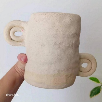 Caneca de cerâmica bege com dois pegadores, fundo branco e planta verde