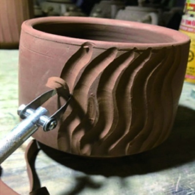 Recipiente cilíndrico de barro vermelho com textura ondulada sendo esculpido com ferramenta metálica.