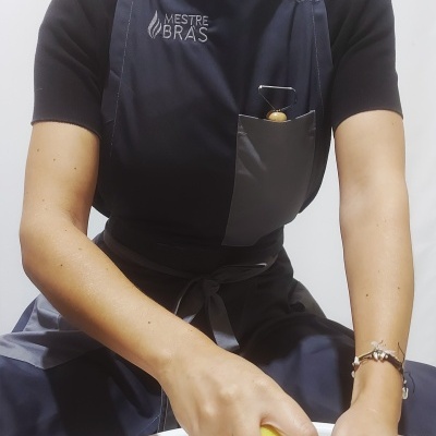 Pessoa com avental azul escuro 'MESTRE BRÁS' segurando esponja amarela sobre bacia branca