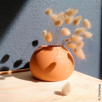 Vaso de barro laranja com flores secas bege claro sobre superfície de madeira clara e parede cinza