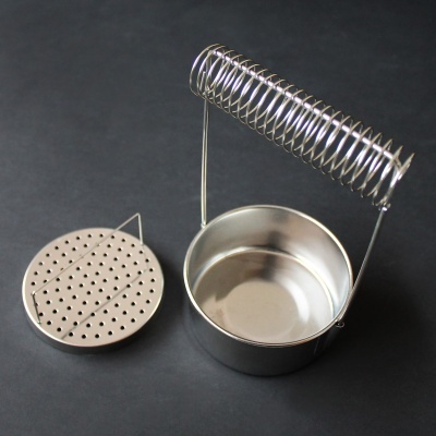 Conjunto metálico prateado com base, peça perfurada e estrutura com mola sobre superfície escura