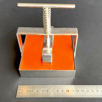 Prensa metálica prateada com superfície laranja e régua de metal em fundo preto