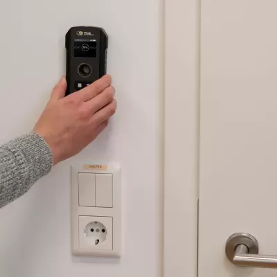 Dispositivo de medição digital preto numa parede branca perto de interruptores e tomada.