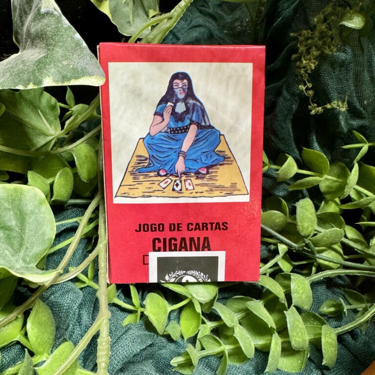 Jogo de Cartas Ciganas Caixa vermelha de baralho Jogo de Cartas Cigana com ilustração de mulher sentada em tapete, entre plantas verdes.