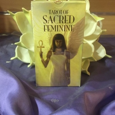 Caixa de tarô TAROT OF SACRED FEMININE com imagem de mulher e asas sobre tecido roxo e flores brancas.