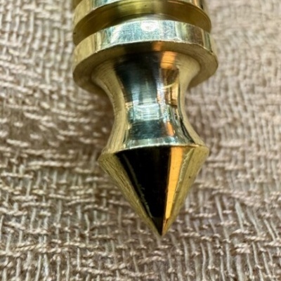 Pêndulo metálico dourado polido com espiral sobre tecido texturizado marrom.