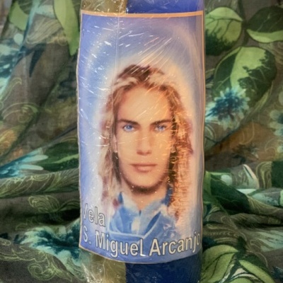 Vela azul e amarela com rótulo de São Miguel Arcanjo sobre tecido verde com padrão de folhas
