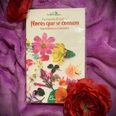 Livro com capa branca e flores coloridas sobre tecido roxo com flores vermelhas artificiais.