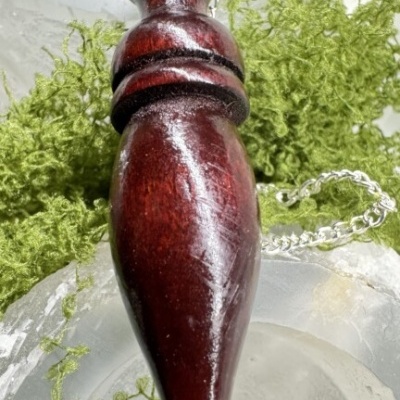 Pingente em madeira castanho escuro com corrente prateada sobre pedras e folhas verdes