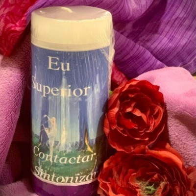Vela branca com rótulo azul e texto em português rodeada por toalhas e flores vermelhas.