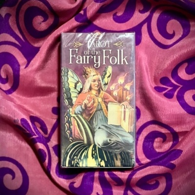 Caixa de tarot com uma fada ilustrada, sobre tecido rosa e roxo.