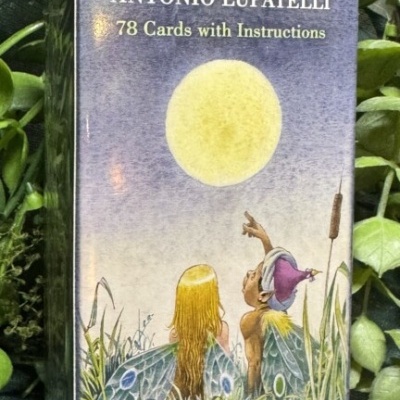 Caixa de baralho de tarot com ilustração de figuras junto a uma grande lua amarela e plantas verdes