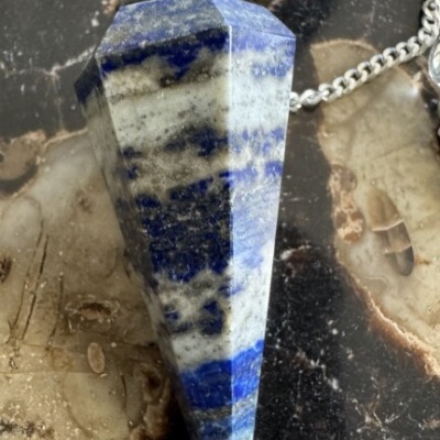 Pendente de pedra azul e branca com corrente prateada sobre fundo de pedra marrom e preto