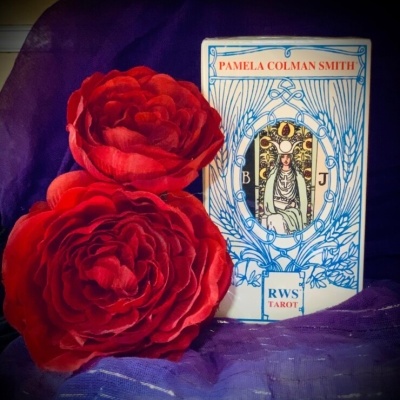 Caixa de cartas de tarot com texto e duas flores vermelhas artificiais junto a tecido roxo.